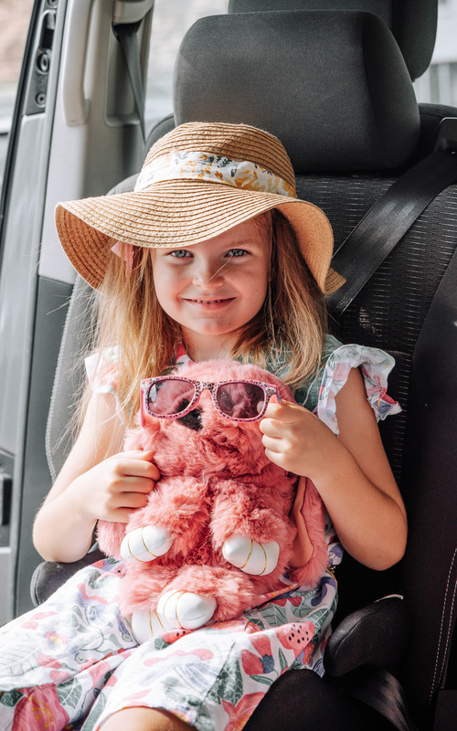 Smiling girl in a sun hat holds a pink stuffed animal with sunglasses while sitting in a car seat.