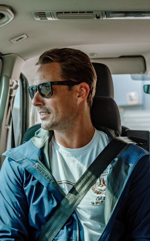 Ein Mann mit Sonnenbrille und Sicherheitsgurt sitzt auf dem Rücksitz eines Autos, schaut aus dem Fenster und lächelt.