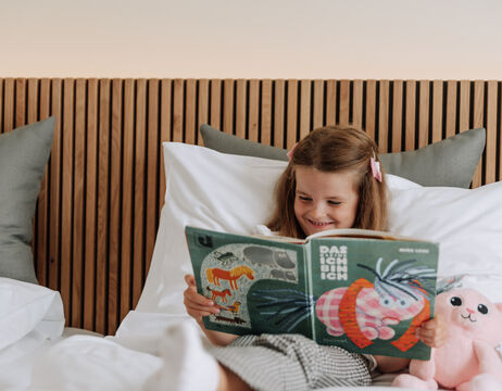 Ein lächelndes Mädchen liest ein Bilderbuch im Bett, mit einem Stofftier und Kissen neben sich.