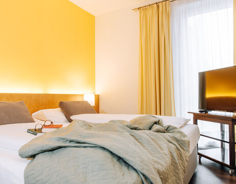 Modernes Hotelzimmer mit einem Doppelbett, grauer Decke, zwei Kissen, TV und Sonnenlicht durch die Vorhänge.