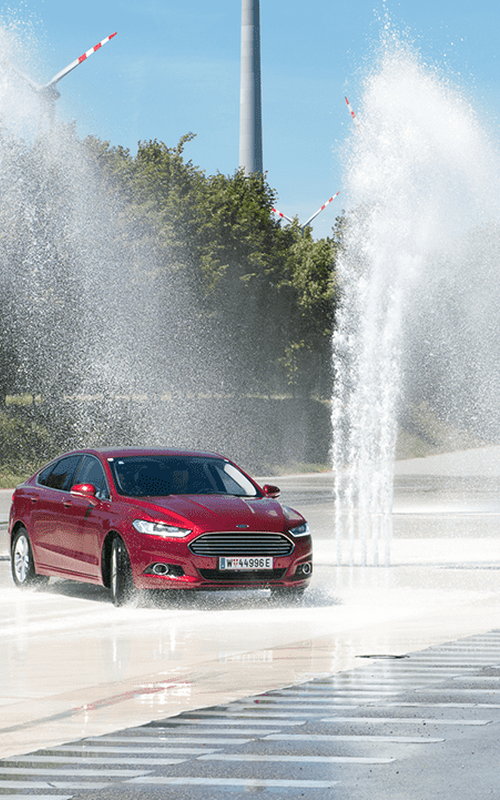 Ein rotes Auto fährt auf einer nassen Straße durch Spritzwasser, im Hintergrund sind Bäume und Windräder zu sehen.