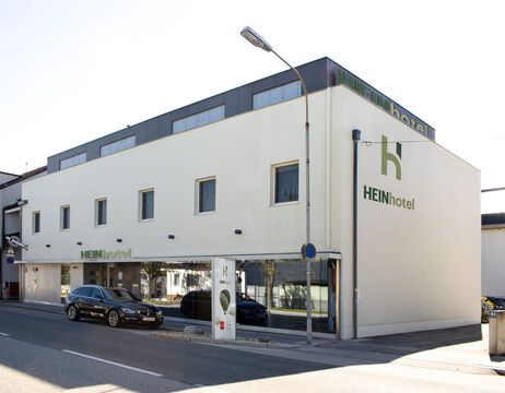 Modernes weißes Gebäude mit HEINhotel-Schriftzug, großen Fenstern und parkenden Autos in einer ruhigen Straße.