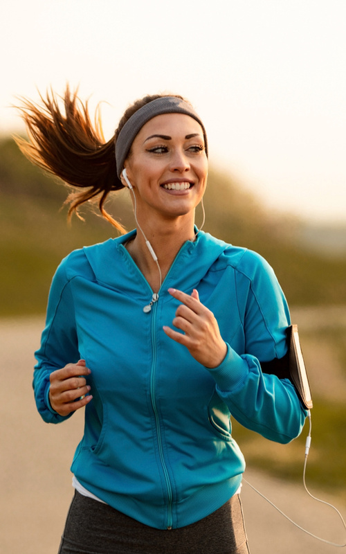 Woman in blue jacket jogging outdoors, smiling, wearing headphones and a headband, with a phone on her arm.