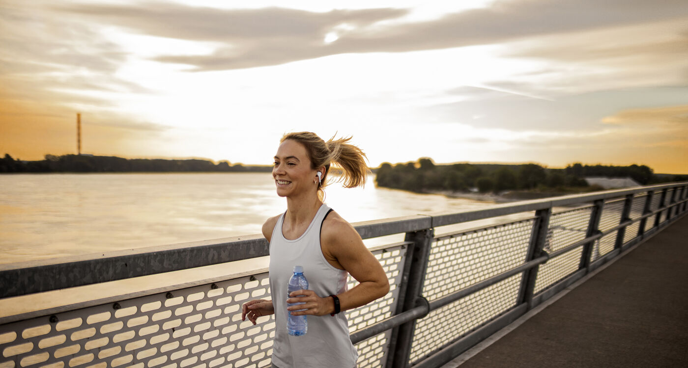 Woman jogging along a riverside path at sunset, holding a water bottle and wearing headphones.