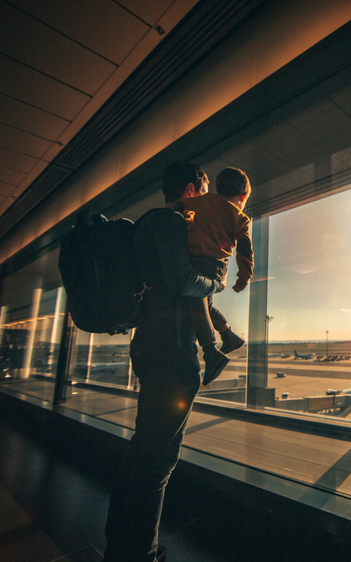 Ein Erwachsener, der ein Kind im Arm hält, schaut aus dem Flughafenfenster auf Flugzeuge bei Sonnenaufgang oder Sonnenuntergang.